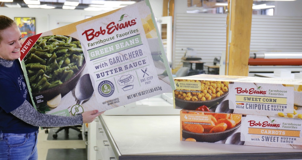 Kaleidoscope team member holding oversized Bob Evans Farmhouse Favorites packaging prototype in production facility